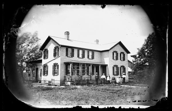 Knut Knudesen family in yard of large brick house with shuttered windows, stone foundation and trellis at porch.