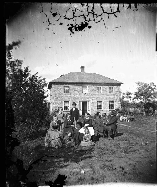 The Sjur Starkson Reque home. From left to right, seated, are Martha Reque, Mari Reque Lee, Brita Reque Quale, Nels Lee and Ole Quale. Standing are Anna Maria Reque and Louis Quale. Sitting on the ground in front are Julia Lee and a daughter of Brita and Ole Quale. Farmhands sit on the grass in the background with a dog.