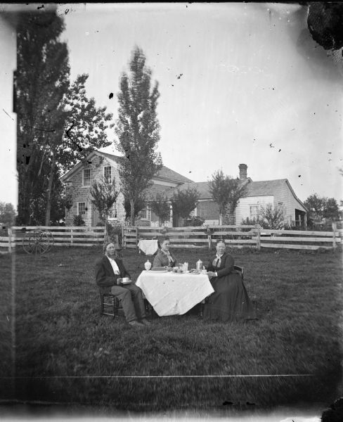 Family seated in yard around set table drinking coffee. Behind them is an upright and wing stone house with a frame addition and latticework and board fencing.