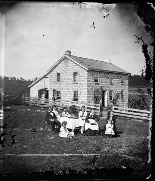 Family seated around table in yard with stone house and frame addition in back; wood fence along side of house.