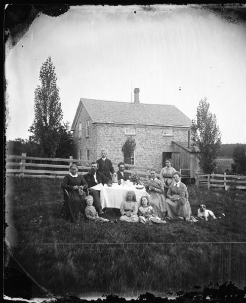 Family around table in yard. Four women, three men, and four children. The woman at far left is knitting, and on the far right is a dog. In the background is a small stone house behind a wood fence. The house has a small frame addition at the front right, probably a kitchen.