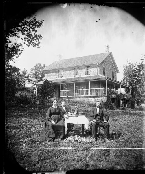Family, including a woman, young man and man sitting around table in a yard, with a two-story stone house with a porch on at least two sides. A young girl is standing behind a railing on the portch. L.E. Johnson was the neighbor of B.F. Halin. The farm was located in Section 19 NW.