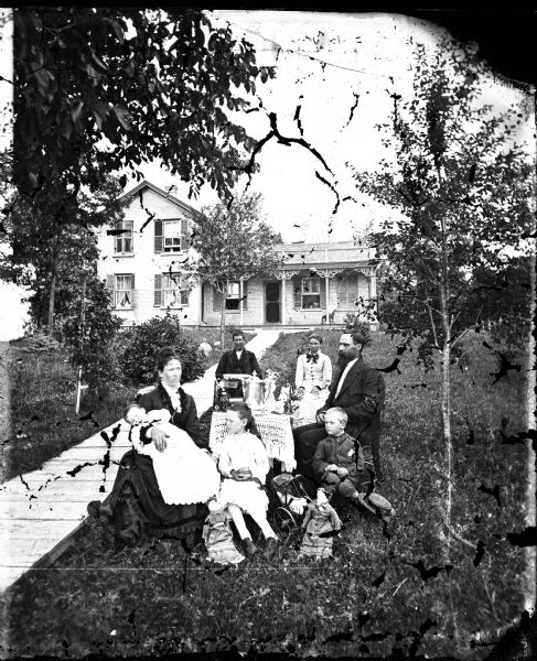 Family in yard around table, with mother holding baby and doll. There is a doll carriage on the ground, and a long plank walkway leads to a brick house with carpenter's lace porch trim and second story shutters.
