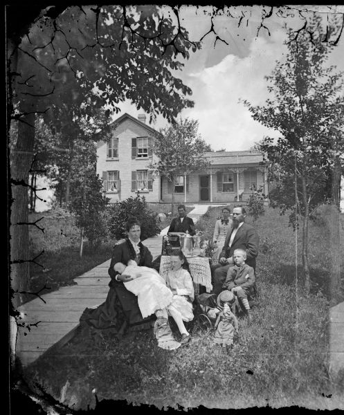 Family in yard around table, with mother holding baby and doll. There is a doll carriage on the ground, and a long plank walkway leads to a brick house with carpenter's lace porch trim and second story shutters closed.