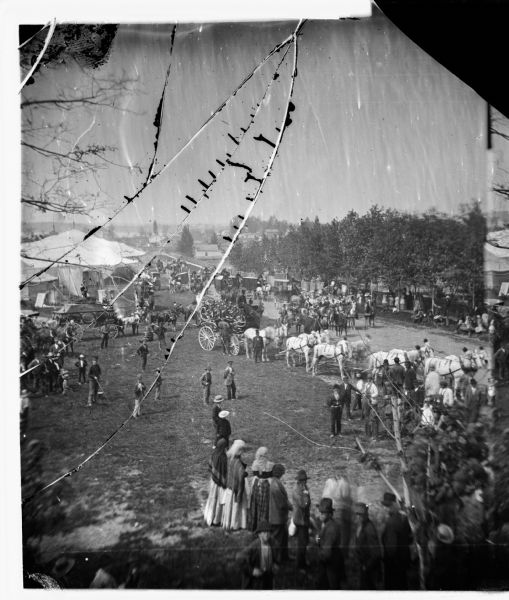 Elevated view of ornate carriage and animal cage with tents and people looking on at the annual Wisconsin State Fair held at Camp Randall.