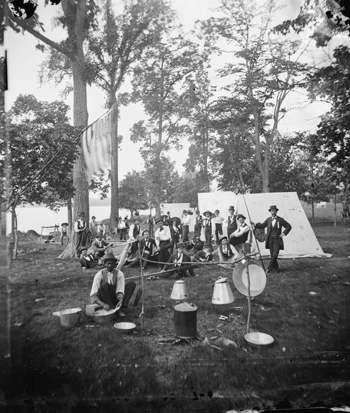 Chicago Traveling Club encamped at McBride's Point at Maple Bluff on Lake Mendota near Madison.  An African American sits at the left foreground with pans and utensils for cooking. One man has a flute and others pose with fishing equipment. A flag hangs from the tree on the left.