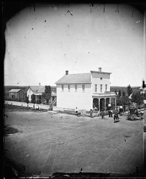 Elevated view of the J. Green & Co. store, a frame building with a false front and a third story fan window. Men with farm implements stand nearby with horses and a carriage. A picket fence surrounds the store and other buildings behind it.