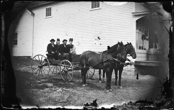 Two couples in a horse-drawn wagon outside of a commercial frame structure, probably a store.