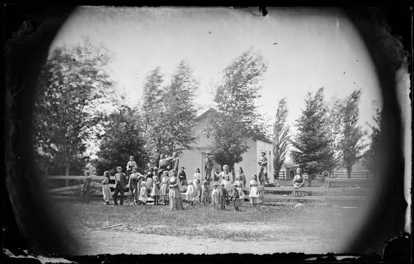 Twenty-nine people (teacher with children) near board fence in front of a one room schoolhouse with outhouse.