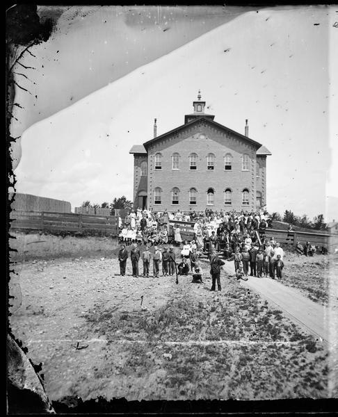 Lodi School was a three-story brick Romanesque building with a small cupola, built in 1868. The school burned on March 28, 1878. It was rebuilt by December 1878, but burned again in 1886. A large crowd of boys, girls and teachers is gathered on plank sidewalks and a plank fence in the foreground.