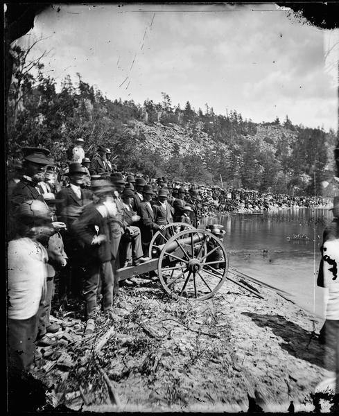 A huge crowd, mostly men, has gathered on the bank of Devil's Lake to witness the firing of a cannon. The occasion is the Grand Regatta held June 21-22, 1877.