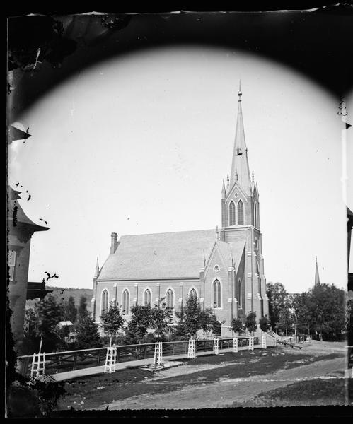 View of the front and side of the First Norwegian Lutheran Church.  Trees along the sidewalk have protective wooden trellises. This view is part of a set of stereographs entitled "The Synod at Decorah, 1876" from Dahl's 1877 "Catalog of Stereoscopic Views."