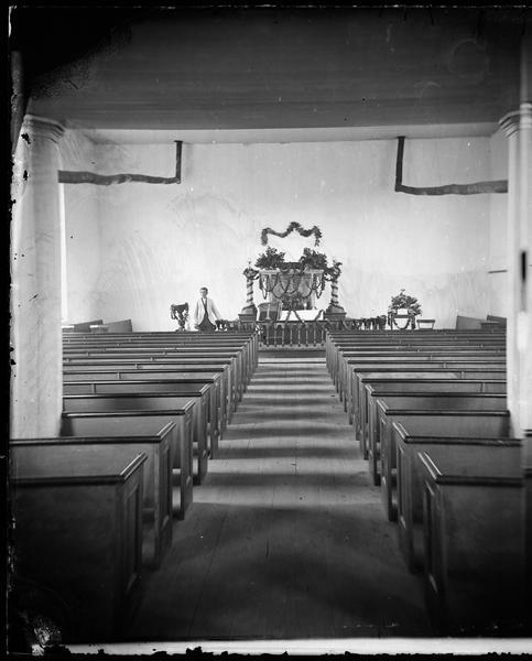 View of church interior looking up the aisle towards an altar decorated with garlands, possibly for a wedding. A young man stands at the left of the altar. Twin stove pipes are running along the back wall just under the ceiling.
