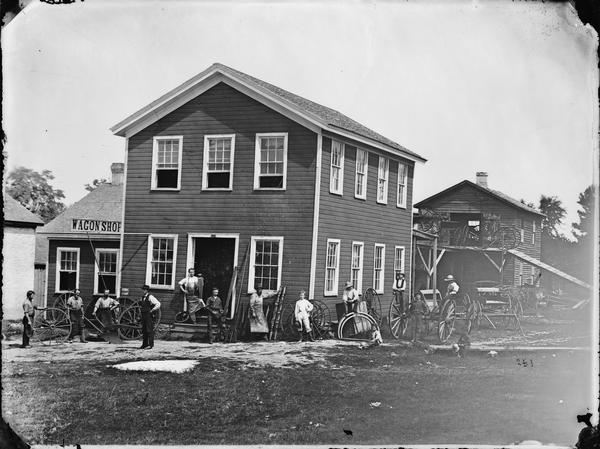Men and boys stand in front of a wagon shop, probably in Edgerton, on Fulton Street. Wagons, wheels, yokes and farm implements surround them. A ramp behind the building was for wagons to go up to the shop's second story.