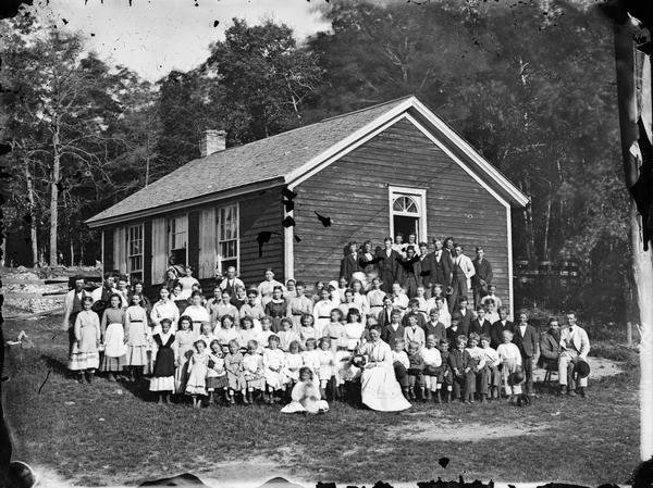 Group portrait of children and teaches posing in front of a small frame school building.