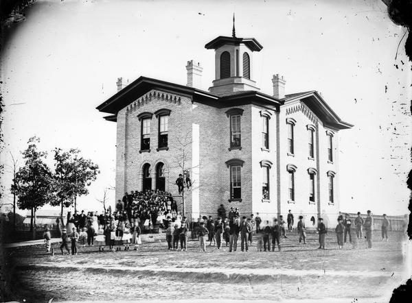 Large brick two-story school house with small tower and three chimneys. A group of students are in front of the building and two boys are in a tree.