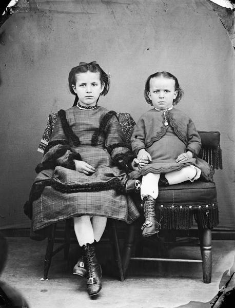 Portrait of two unidentified children holding hands while posing on homemade fancy chairs. The girl is dressed in a hand-me-down dress cut from a heavy wool woman's dress, while the boy on the right is wearing an old-fashioned infant's dress which has become too small for him.