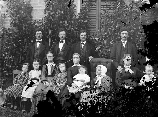 Extended family of fourteen posed in their best clothes outside a home. Older woman seated in rocking chair, with men, women, girls, boy and baby.