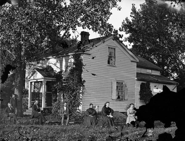 Newhouse family in yard under large tree, with frame house that has latticework siding on the front porch as well as a Hekla sign, indicating that the house is insured by the Hekla Fire Insurance Company. Woman on far right holds a book. They are from left to right: Knute Newhouse (seated), unknown (leaning against tree), Ole Newhouse, Christian Newhouse,  Kari Newhouse, Anna Newhouse, and Rachel Larson.