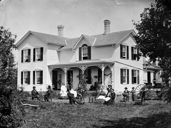 This is said to be the family of Hans Holtan (a.k.a. Austinson) posed in the yard before a frame house that has carpenter's lace on porch, two separate doors, shutters and a fan shape above window on gable. Numerous house plants are on the porch and a man sits in an unusual bucket chair holding out what appears to be a newspaper. In the 1870 census the family consisted of Hans 36, Rana 37, Austin 9, Jergen 1, Jane 5, John 7 and Ole 4 1/2. Holtan's sister Rachel 38 also lived there as well as two male farm laborers.