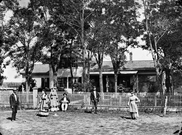 Six people playing croquet in front of picket fence; frame house with shutters and latticework on porch in background.