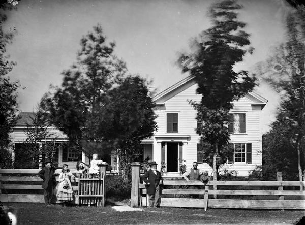 Family by wood fence with one child sitting on top of gate; wood house with upper window shutter closed and shutter trim around doorway; trellis by porch. A hand saw is leaning against the fence.