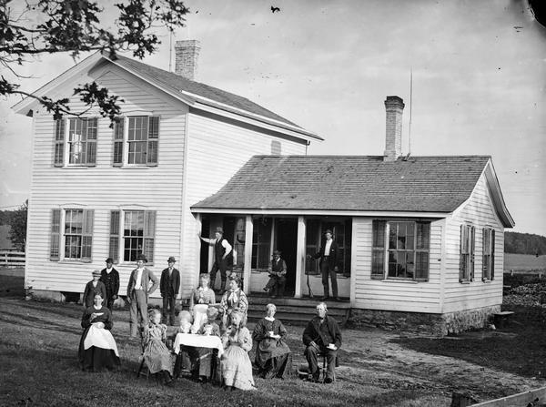 A family of sixteen in yard around table set for coffee. Three men are in the background, one holding a shotgun. There is a frame house with shutters and stone foundation behind them.