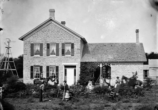Group portrait of family in yard. Two women are posing on horses, one man has a rifle, and another man has a hoe. A woman is sitting in front of the porch with a child in her lap. Behind the group is the stone house with a frame building in the right background, and a windmill at the side of the house on the left.