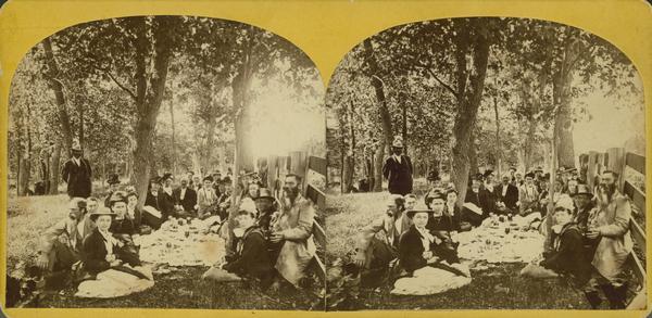 "The Grand Regatta, at Devil's Lake, June 21st & 22nd, 1877." A  crowd sits on the ground about to commence lunch at Devil's Lake.