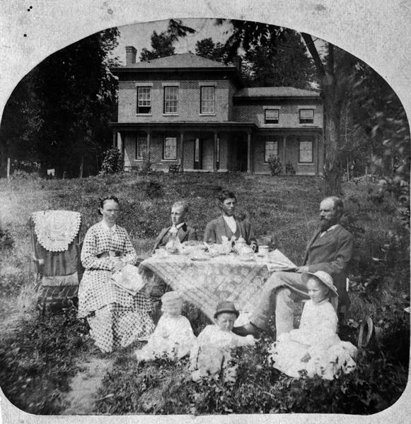 Halle Steensland and his wife Sophia pose with their five children at the family home, later located at 733 Lakewood Boulevard, Village of Maple Bluff. (The house and farm were later owned by Robert M. La Follette, Sr.) The two older boys, Halbert and Henry, sit behind a table set for coffee and covered with a beautiful lace table cloth. On the ground before the table sit the younger children, Edward, Morton and Helen. A carriage and outbuilding, probably a carriage house, are visible.
