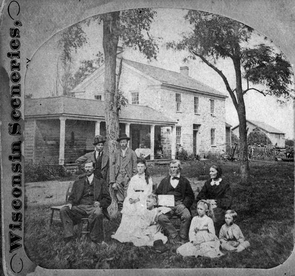 Half of a stereograph showing a family of nine sitting in front of a stone two-story house. Behind it on the right are various horse-drawn farm implements, a large circular haystack and an outbuilding. This is possibly the Springen family.