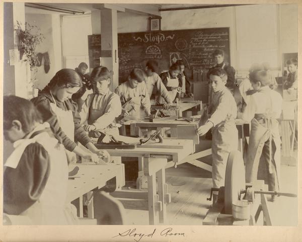 Students working in a sloyd room at the Indian Industrial School.  A sloyd is a type of wood-carving knife.
