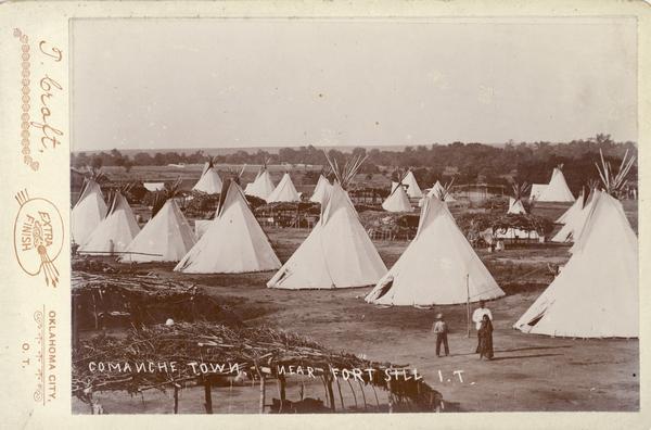 Tepees in a Comanche town near Fort Sill, Indian Territory. A woman and two men gather in the foreground.