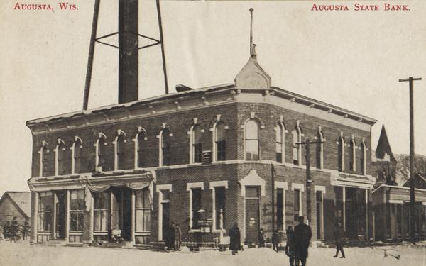 View from street of the Augusta State Bank on a corner. There are pedestrians walking on the street and along the sidewalk. There is a support structure for what may be a water tower rising behind the bank building. Caption reads: "Augusta, Wis. Augusta State Bank."