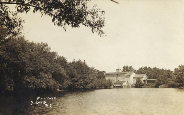 Caption reads: "Mill Pond, Augusta, Wis." View across water towards a large building on the far shoreline. Tree-lined shore on the left.