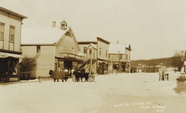 View of a main street during the winter. One horse is covered with a horse blanket. Caption reads: "Winter Scene at Blue Mounds Wis."