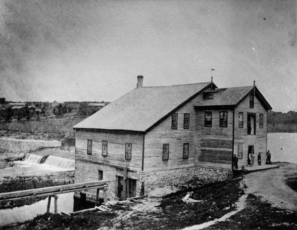 View of the Rockdale mill, a grist and sawmill built in 1847 by Thomas and Nathan Van Horn. A group of people are standing on a sidewalk or loading dock on the right.