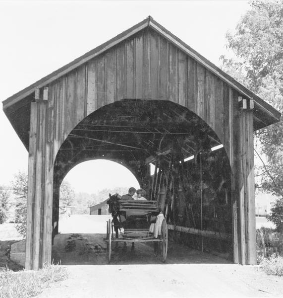 Stonefield Village Rear view of a couple sitting in a horse-drawn buggy which is driving through a covered bridge at Stonefield Village. Buildings and trees are in the distance.