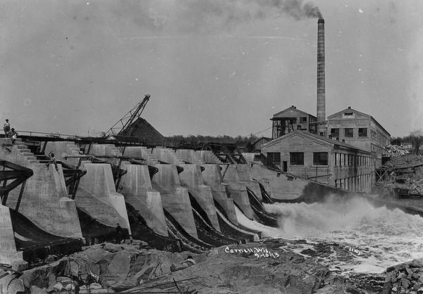 Dam and hydroelectric power house, with a billowing smokestack and a crane in the background.