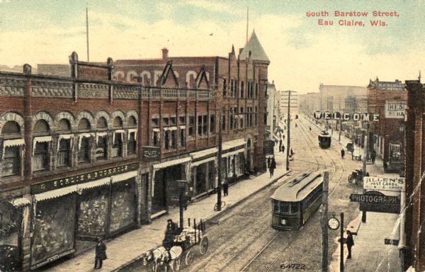 Elevated view of a main throughfaire, with buildings lining each side of the street, with many pedestrians, a horse-drawn carriage, a clock hanging from a pole in the street, and trolleys running along lines.  There is also a sign on a building that reads: "Allen's Post Cards."  Another sign reads: "Isaac's Photography." A "Welcome" sign hangs from a line over the street. Caption reads: "South Barstow Street, Eau Claire, Wis."
