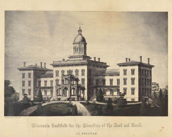 Engraved view of the building and the grounds. On the top of the dome is a wind vane. Pedestrians, an equestrian, and a man in a horse-drawn carriage are in the foreground. Caption reads: "Wisconsin Institute for the Education of the Deaf and Dumb. At Delavan."