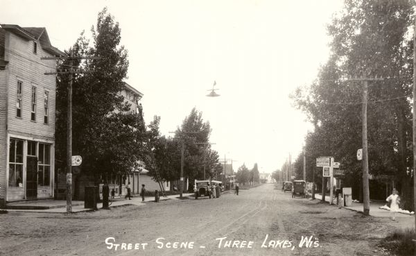 View down unpaved street. Caption reads: "Street Scene — Three Lakes, Wis."