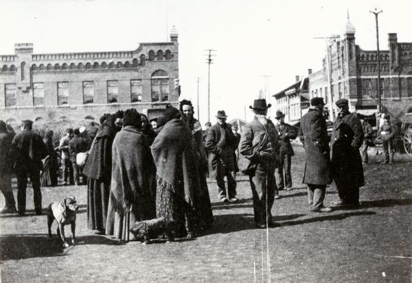 Polish immigrants in Market Square wearing Polish national dress. The men are wearing suits, coats, and hats. The women are wearing dresses, shawls and hats, and two dogs stand near them. Large brick buildings are in the background.