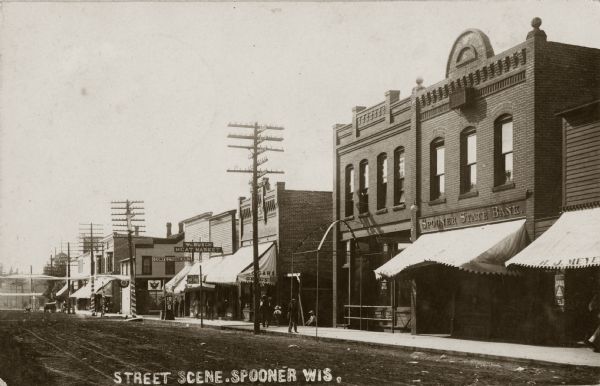 View of a street scene, with the Spooner State Bank in the foreground, and the William Busch Meat Market and a confectionary further down the street. Caption reads: "Street Scene, Spooner Wis."