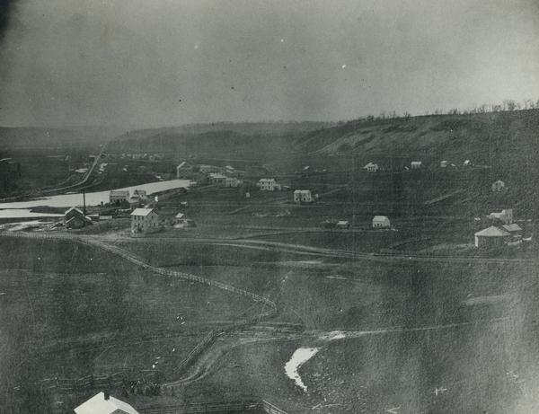Elevated view of multiple buildings in Cross Plains from Lutheran Church Hill. Many roads and fences cross the fields. Wooded hills are in the background. In the lower left people are gathered near a building.