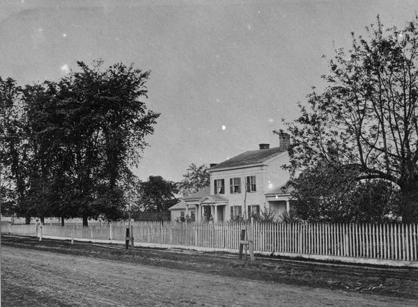 View across unpaved road towards Randall Wilcox's home, with a fence along the sidewalk. A group of people are standing on the front porch.