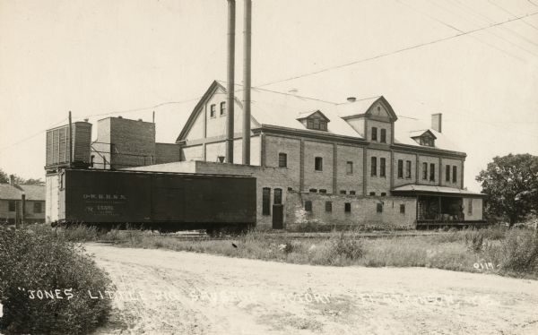 View of Jones' sausage factory with train car on railroad tracks in front. Caption reads: "'Jones Little Pig Sausage Factory' Ft. Atkinson, Wis."