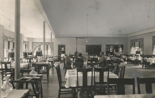 Interior of the Sherwood Forest Hotel dining room.  Men and women are seated at a table for a meal, and the uniformed waitstaff stand to the left.