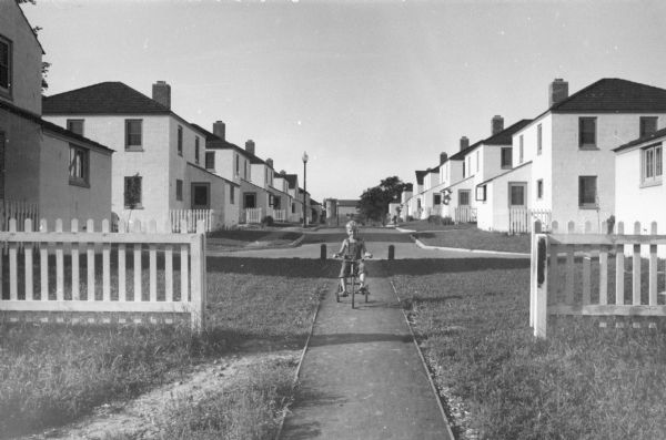 A child peddles his tricycle on a sidewalk at the end of a cul de sac in his Greendale neighborhood with houses lining the street.