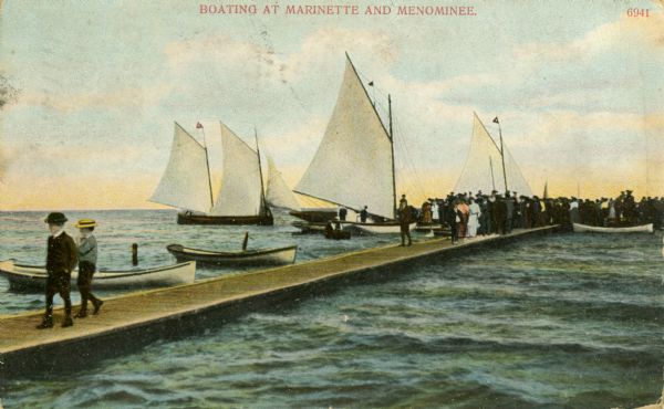 View of a boating scene off of the shore of Marinette and Menominee. Two boys are walking on the pier on the left, and a crowd of people are gathered at the end of the pier near sailboats. Caption reads: "Boating at Marinette and Menominee."